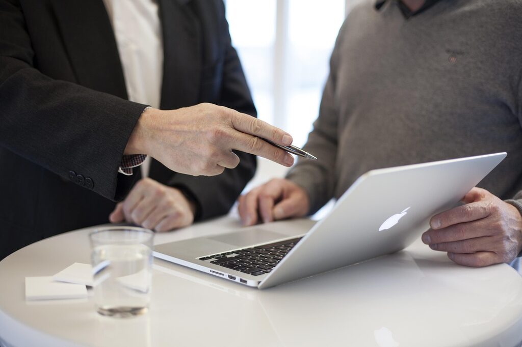 Illustration of a New Zealand small business owner and a web designer talking across a table with a laptop, surrounded by labels for experience, process, timeline, pricing and support — the ten questions to ask before hiring