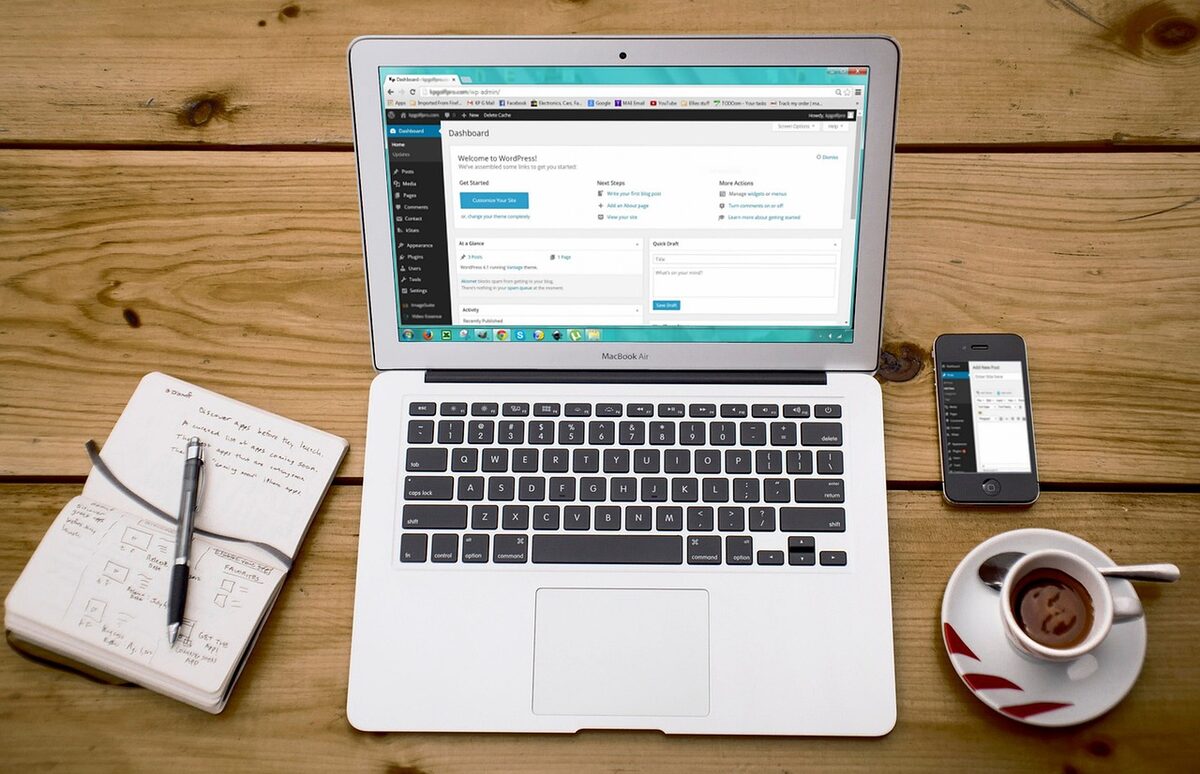 Laptop showing a website dashboard on a warm wooden desk beside a notebook with hand-drawn website wireframe sketches